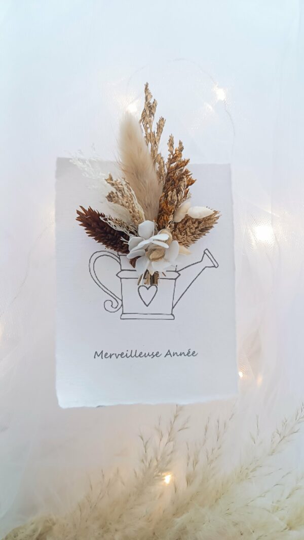 Carte de vœux artisanale “Merveilleuse Année” présentée dans une mise en scène poétique. Sur un fond doux et lumineux, la carte blanche est décorée d’un petit bouquet de fleurs séchées naturelles — pampas, blé, gypsophile et feuillages dorés — fixées au-dessus d’une illustration d’arrosoir avec un cœur. Elle repose sur une rondelle de bois entourée de plumes de pampa et de guirlandes lumineuses, créant une ambiance chaleureuse et naturelle. Carte de remerciement pour cette nouvelle année - carte à offrir personnalisé pour noël et bonne année -