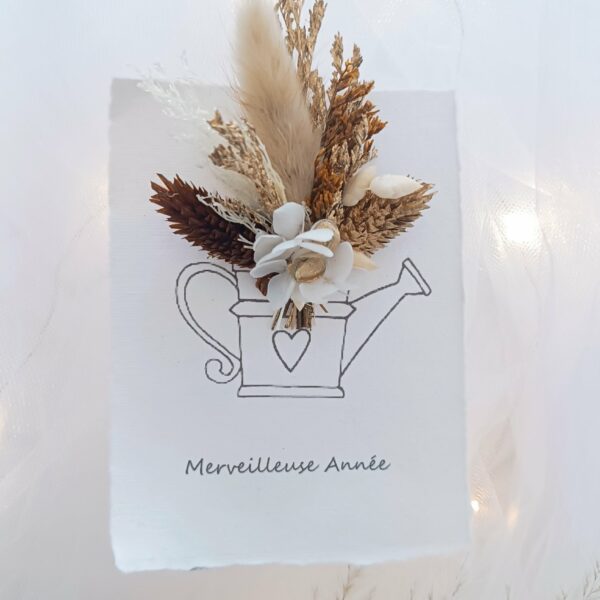 Carte de vœux artisanale “Merveilleuse Année” présentée dans une mise en scène poétique. Sur un fond doux et lumineux, la carte blanche est décorée d’un petit bouquet de fleurs séchées naturelles — pampas, blé, gypsophile et feuillages dorés — fixées au-dessus d’une illustration d’arrosoir avec un cœur. Elle repose sur une rondelle de bois entourée de plumes de pampa et de guirlandes lumineuses, créant une ambiance chaleureuse et naturelle. Carte de remerciement pour cette nouvelle année - carte à offrir personnalisé pour noël et bonne année -