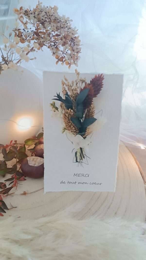 Carte de vœux artisanale “Merveilleuse Année” présentée dans une mise en scène poétique. Sur un fond doux et lumineux, la carte blanche est décorée d’un petit bouquet de fleurs séchées naturelles — pampas, blé, gypsophile et feuillages dorés — fixées au-dessus d’une illustration d’arrosoir avec un cœur. Elle repose sur une rondelle de bois entourée de plumes de pampa et de guirlandes lumineuses, créant une ambiance chaleureuse et naturelle. Carte de remerciement pour cette nouvelle année - carte à offrir personnalisé pour noël et bonne année -