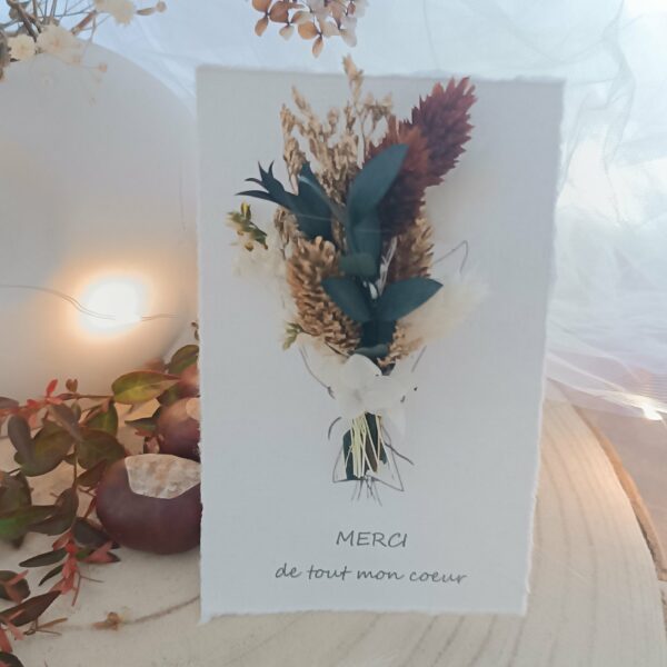 Carte de vœux artisanale “Merveilleuse Année” présentée dans une mise en scène poétique. Sur un fond doux et lumineux, la carte blanche est décorée d’un petit bouquet de fleurs séchées naturelles — pampas, blé, gypsophile et feuillages dorés — fixées au-dessus d’une illustration d’arrosoir avec un cœur. Elle repose sur une rondelle de bois entourée de plumes de pampa et de guirlandes lumineuses, créant une ambiance chaleureuse et naturelle. Carte de remerciement pour cette nouvelle année - carte à offrir personnalisé pour noël et bonne année -