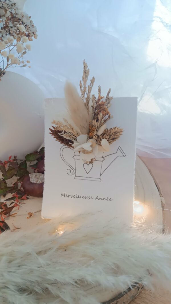 Carte de vœux artisanale “Merveilleuse Année” présentée dans une mise en scène poétique. Sur un fond doux et lumineux, la carte blanche est décorée d’un petit bouquet de fleurs séchées naturelles — pampas, blé, gypsophile et feuillages dorés — fixées au-dessus d’une illustration d’arrosoir avec un cœur. Elle repose sur une rondelle de bois entourée de plumes de pampa et de guirlandes lumineuses, créant une ambiance chaleureuse et naturelle. Carte de remerciement pour cette nouvelle année - carte à offrir personnalisé pour noël et bonne année -