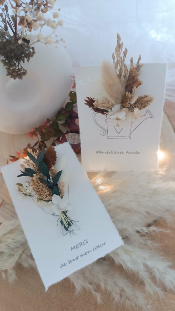 Carte de vœux artisanale “Merveilleuse Année” présentée dans une mise en scène poétique. Sur un fond doux et lumineux, la carte blanche est décorée d’un petit bouquet de fleurs séchées naturelles — pampas, blé, gypsophile et feuillages dorés — fixées au-dessus d’une illustration d’arrosoir avec un cœur. Elle repose sur une rondelle de bois entourée de plumes de pampa et de guirlandes lumineuses, créant une ambiance chaleureuse et naturelle. Carte de remerciement pour cette nouvelle année - carte à offrir personnalisé pour noël et bonne année -