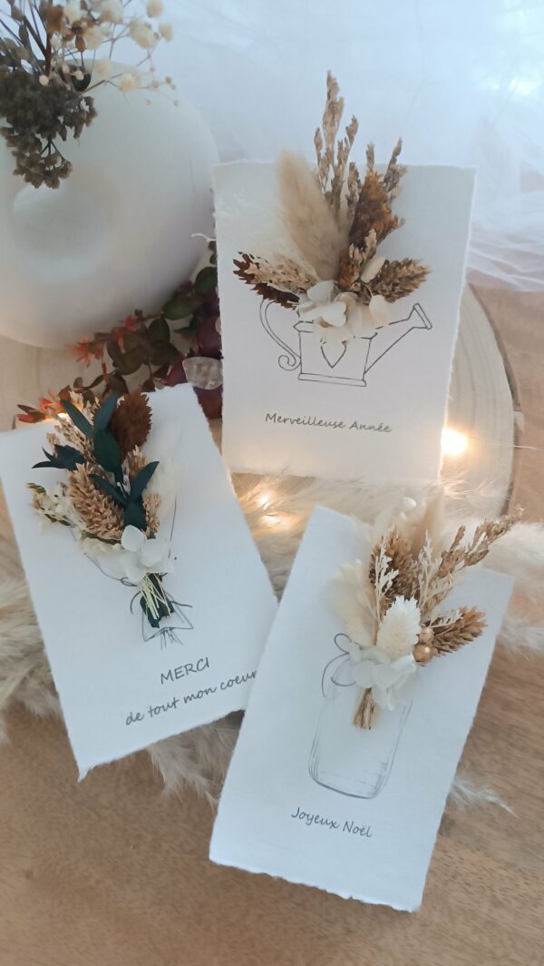 Carte de vœux artisanale “Merveilleuse Année” présentée dans une mise en scène poétique. Sur un fond doux et lumineux, la carte blanche est décorée d’un petit bouquet de fleurs séchées naturelles — pampas, blé, gypsophile et feuillages dorés — fixées au-dessus d’une illustration d’arrosoir avec un cœur. Elle repose sur une rondelle de bois entourée de plumes de pampa et de guirlandes lumineuses, créant une ambiance chaleureuse et naturelle. Carte de remerciement pour cette nouvelle année - carte à offrir personnalisé pour noël et bonne année -