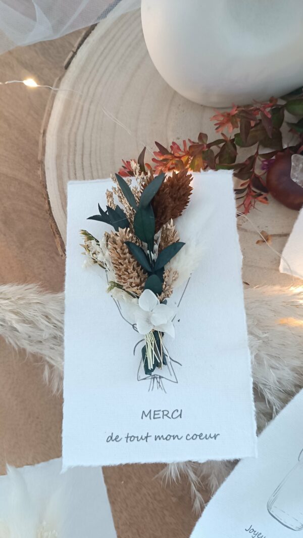 Carte de vœux artisanale “Merveilleuse Année” présentée dans une mise en scène poétique. Sur un fond doux et lumineux, la carte blanche est décorée d’un petit bouquet de fleurs séchées naturelles — pampas, blé, gypsophile et feuillages dorés — fixées au-dessus d’une illustration d’arrosoir avec un cœur. Elle repose sur une rondelle de bois entourée de plumes de pampa et de guirlandes lumineuses, créant une ambiance chaleureuse et naturelle. Carte de remerciement pour cette nouvelle année - carte à offrir personnalisé pour noël et bonne année -