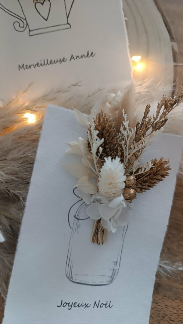 Carte de vœux artisanale “Merveilleuse Année” présentée dans une mise en scène poétique. Sur un fond doux et lumineux, la carte blanche est décorée d’un petit bouquet de fleurs séchées naturelles — pampas, blé, gypsophile et feuillages dorés — fixées au-dessus d’une illustration d’arrosoir avec un cœur. Elle repose sur une rondelle de bois entourée de plumes de pampa et de guirlandes lumineuses, créant une ambiance chaleureuse et naturelle. Carte de remerciement pour cette nouvelle année - carte à offrir personnalisé pour noël et bonne année -