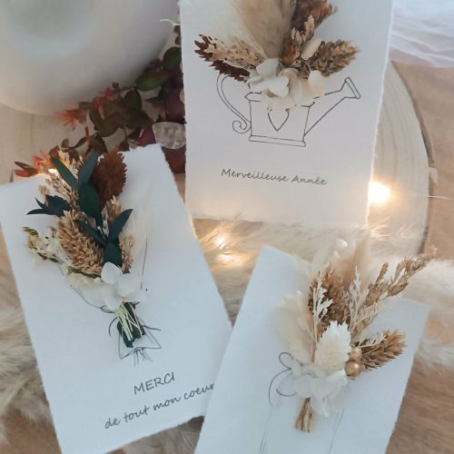 Carte de vœux artisanale “Merveilleuse Année” présentée dans une mise en scène poétique. Sur un fond doux et lumineux, la carte blanche est décorée d’un petit bouquet de fleurs séchées naturelles — pampas, blé, gypsophile et feuillages dorés — fixées au-dessus d’une illustration d’arrosoir avec un cœur. Elle repose sur une rondelle de bois entourée de plumes de pampa et de guirlandes lumineuses, créant une ambiance chaleureuse et naturelle. Carte de remerciement pour cette nouvelle année - carte à offrir personnalisé pour noël et bonne année -